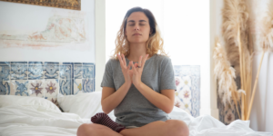 a woman sitting cross-legged on a bed with her eyes closed in a form of meditation pose as part of a summer morning routine