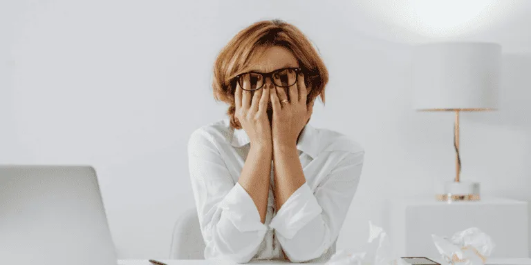 Woman in White Long Sleeve Shirt Wearing Eyeglasses Sitting at a Table with her head in her hands appearing stressed analysing the 3 types of perfectionism