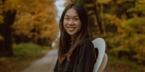 Smiling Woman Standing on a Dirt Road surrounded by trees with amber leaves to symbolise autumn affirmations