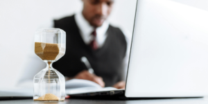 Man in Black Suit Jacket writing in a journal with a lapotp open and a sand timer next to him to symbolise end-of-year journal prompts