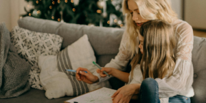 woman writing on a piece of paper with a child in front of the christmas tree - december journal prompts