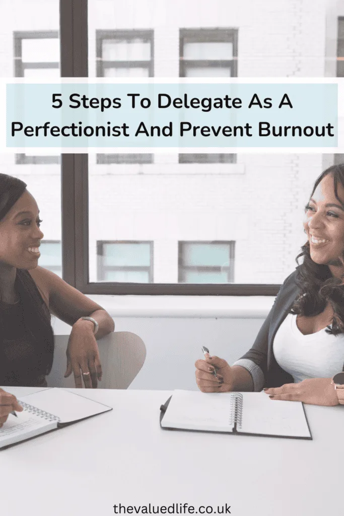 Two professional women smiling and having a discussion at a table, both holding pens and open notebooks. The setting suggests a collaborative work environment, emphasizing the importance of communication and planning when learning to delegate as a perfectionist.