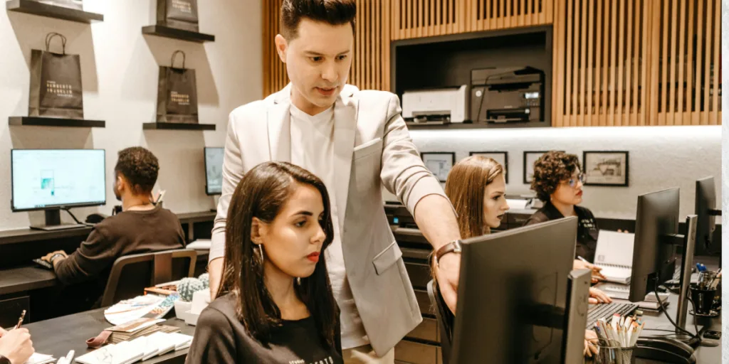 An office scene with a manager pointing at a monitor, while a team member looks intently, highlighting a lack of trust or support in delegation