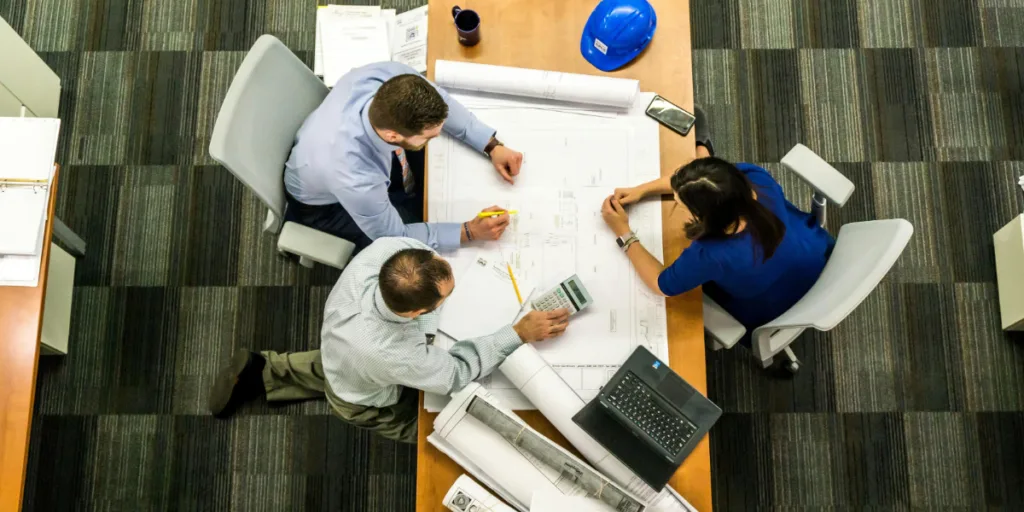 Top-down view of three people at a desk reviewing large blueprints and documents, symbolising micromanagement in project delegation