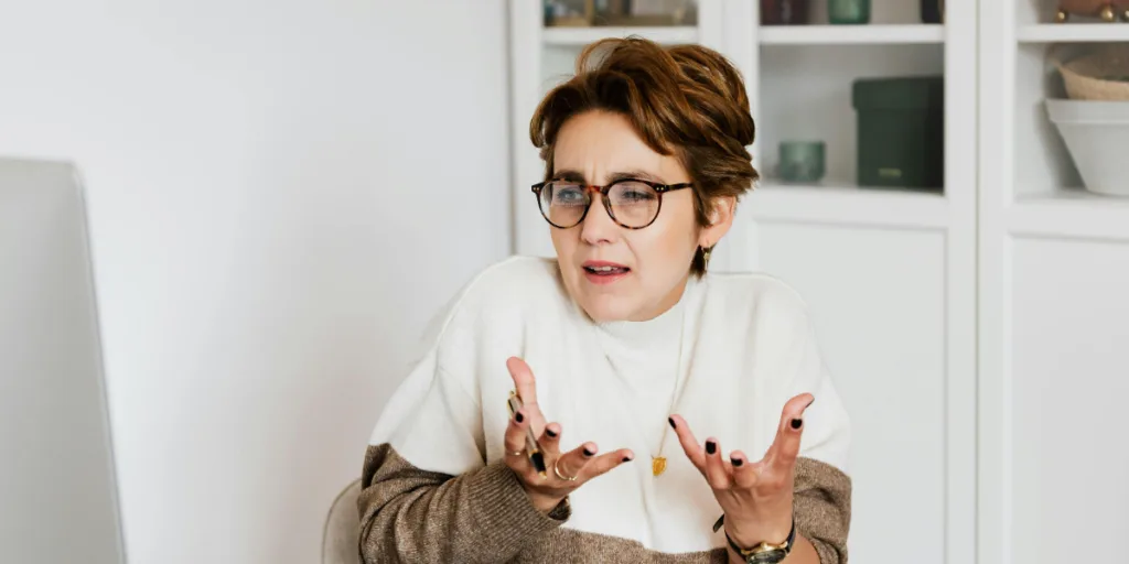 A confused woman wearing glasses and a sweater shrugs her hands in frustration, illustrating the impact of unclear instructions during delegation.
