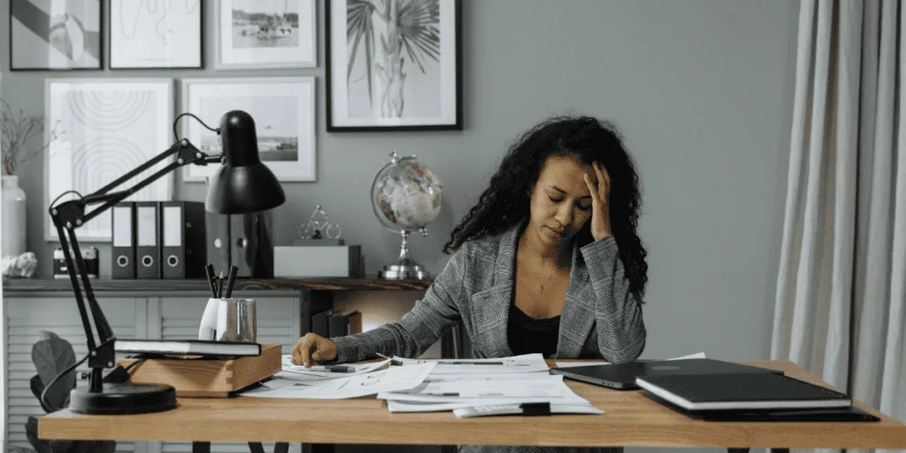 A woman sitting at a desk with her hand on her forehead, appearing stressed while surrounded by papers and a tablet. The office setting and her expression suggest feeling overwhelmed with tasks, highlighting the challenge of learning to delegate as a perfectionist to manage workloads and reduce stress.