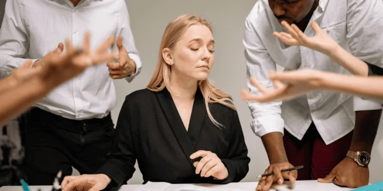 A woman sitting at a desk with her eyes closed, surrounded by colleagues who are gesturing and discussing. She appears overwhelmed by the multiple conversations happening at once. This image illustrates delegation mistakes, such as lack of clear communication or trying to manage every detail, which can lead to confusion and stress for both managers and teams.