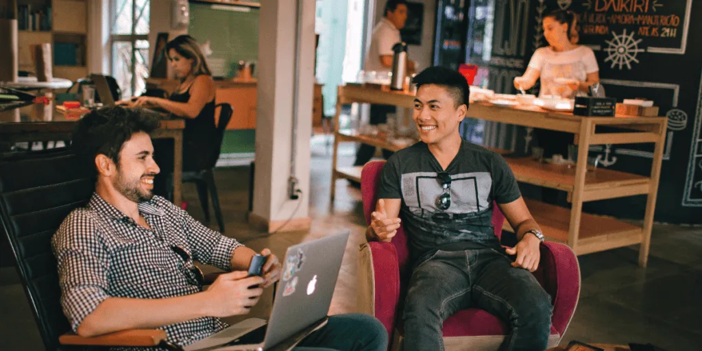 Two men sitting in a casual workspace, smiling and engaging in a relaxed conversation. One holds a smartphone while the other gestures as they chat, suggesting a collaborative environment. The scene emphasizes the importance of open dialogue and teamwork when learning to delegate as a perfectionist