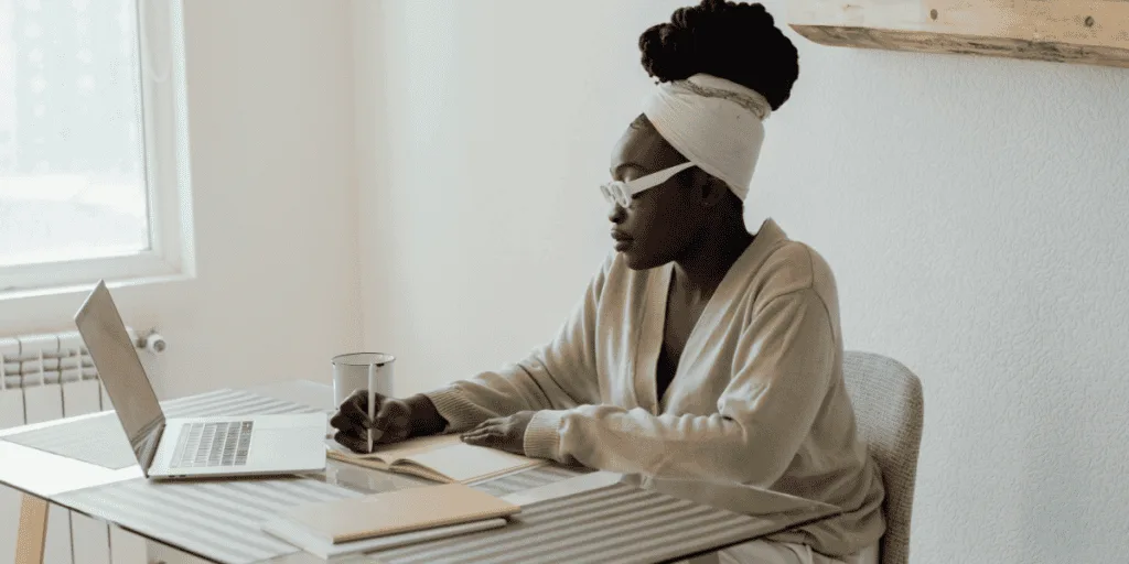 A woman sitting at a desk, focused on writing in a notebook while working on a laptop. She appears reflective and engaged in planning or organizing her thoughts, suggesting the use of journaling or structured note-taking as a tool to delegate effectively as a perfectionist.