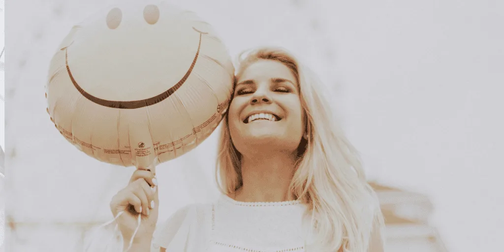 A woman smiling joyfully while holding a yellow balloon with a smiley face, symbolizing the importance of celebrating small successes and embracing positivity in the journey towards healthy striving.