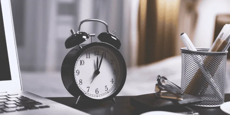 Close-up of a black alarm clock showing 10:10 on a desk next to a laptop and a pen holder, symbolizing productivity and time management myths.