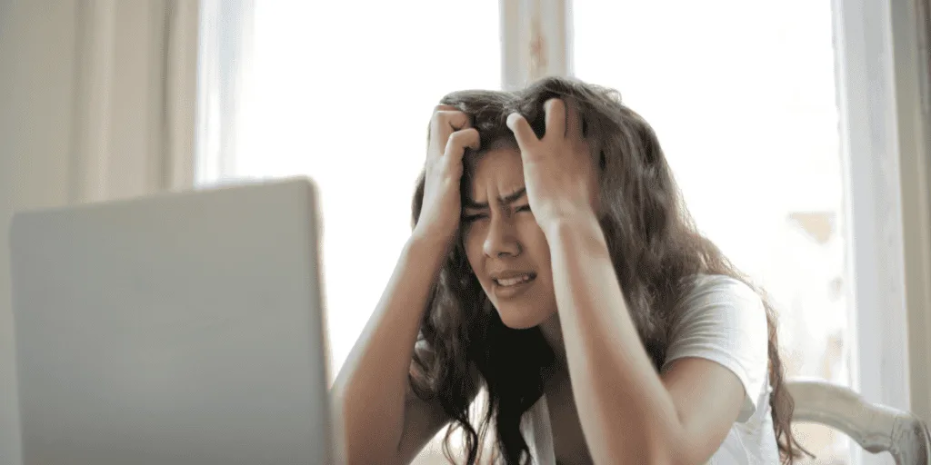 A frustrated woman gripping her head while staring at a laptop, highlighting the stress and frustration that perfectionism can bring, counter to the balanced approach of healthy striving.