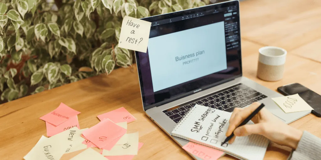 An open laptop with sticky notes and a notepad featuring business planning items, emphasizing the importance of setting realistic goals and focusing on essential actions for a healthy striving mindset
