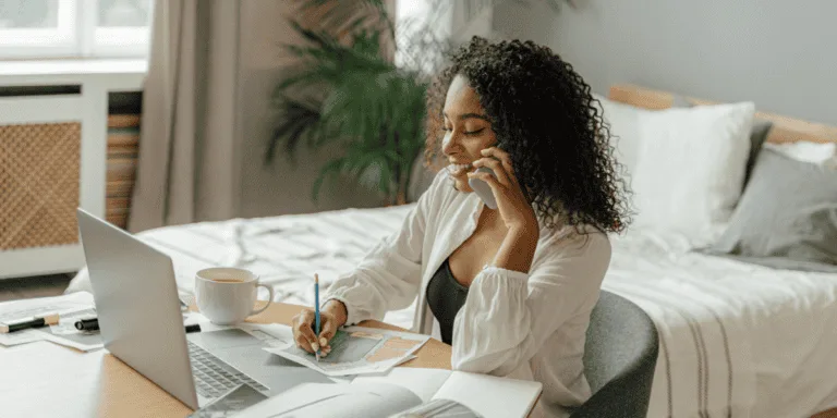 A woman sitting at a desk, planning her week with a notebook, colored markers, and a laptop, while speaking on the phone. This relaxed setting showcases a productive weekly planning routine designed to improve organization and efficiency.