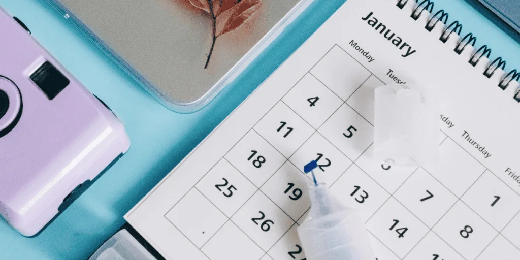 A desk calendar marked with dates alongside a camera and correction fluid, symbolizing the importance of maintaining consistent effort over time to achieve healthy striving without overwhelming oneself