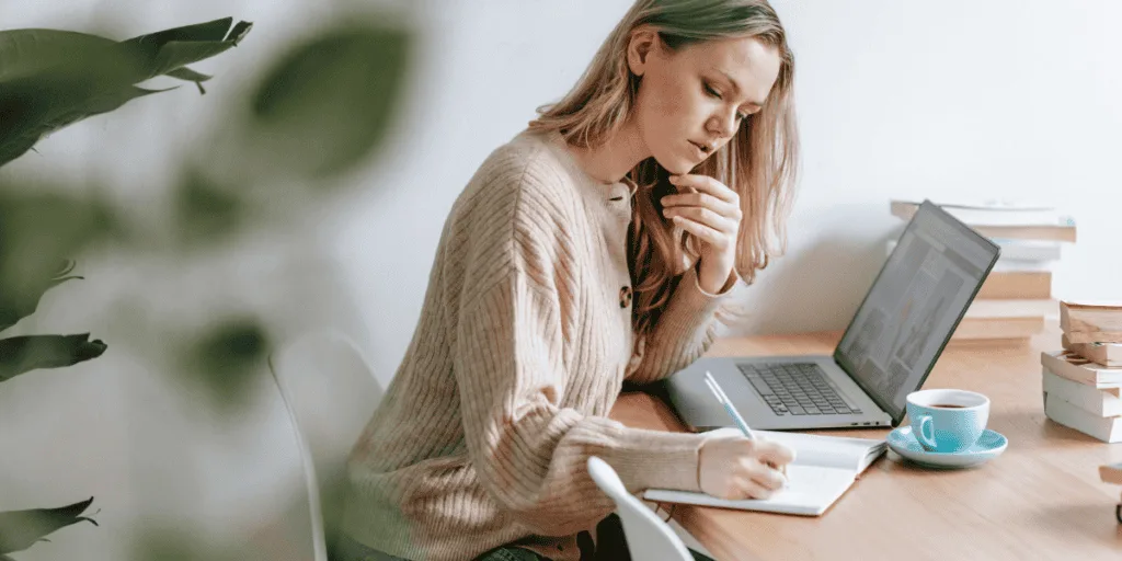 A woman in a cozy sweater sitting at a wooden desk, thoughtfully planning her week in a notebook with a laptop open beside her. This serene scene highlights the benefits of a focused weekly planning routine.