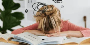 Person with head down on open books, surrounded by scribbled thought bubbles and frowning faces, visually depicting the frustration of being trapped in the perfectionism-procrastination loop