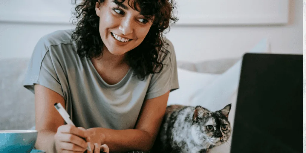 A smiling woman working on her weekly planning routine alongside her cat, with a laptop and pen in hand. This image emphasizes the flexibility of planning in a comfortable home environment.
