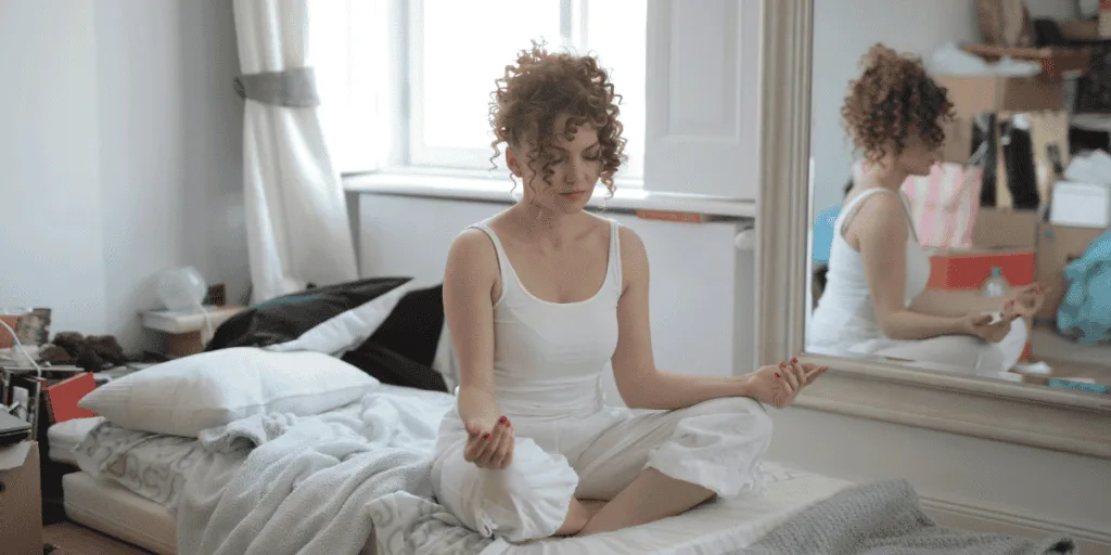 A woman sitting cross-legged on her bed, practicing meditation, representing the role of mindfulness and calm reflection as a core component of a healthy striving mindset