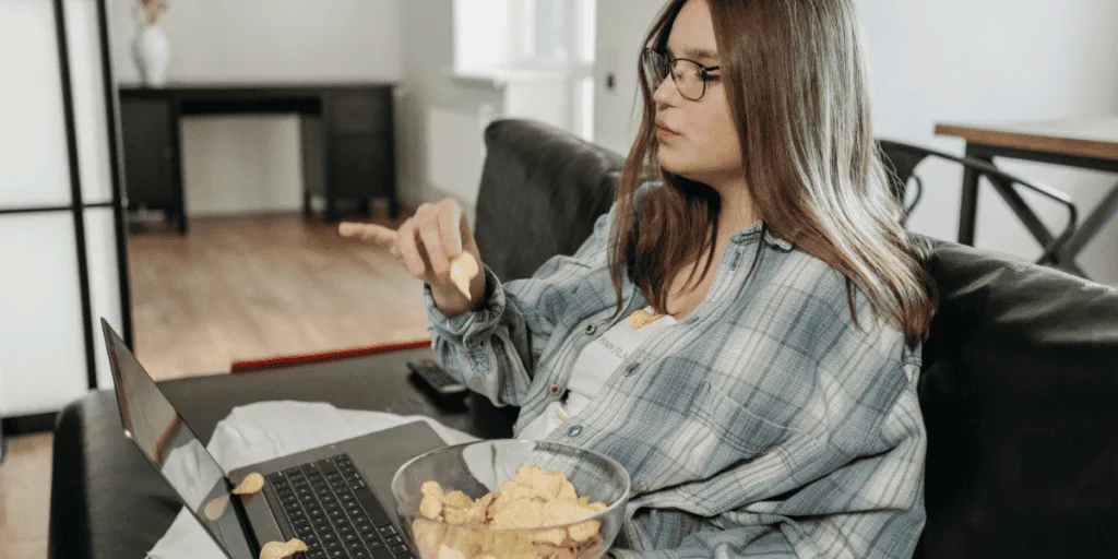 Young woman lounging on a couch with a laptop and a bowl of chips, symbolizing the blurred line between procrastination and laziness, often associated with the lazy perfectionist mindset