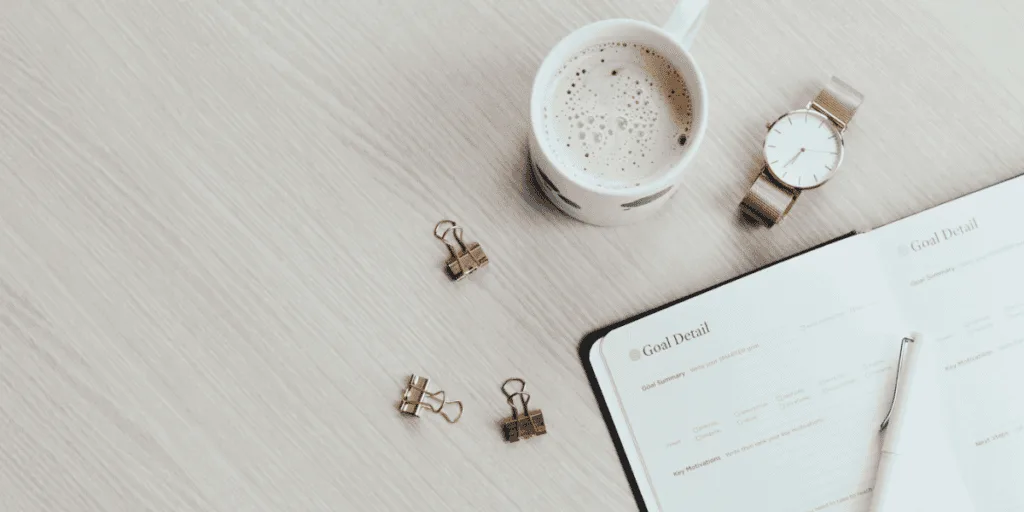 A flat-lay view of a planning setup with a goal-setting notebook, a cup of coffee, a gold watch, and binder clips on a light wooden table. This minimalist scene showcases the tools for an effective planning routine.