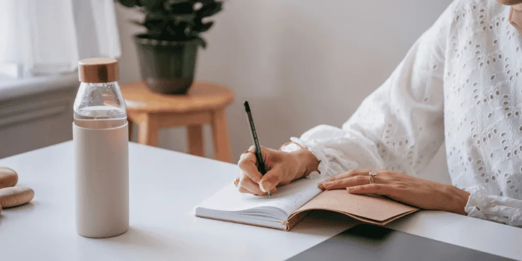 A woman in a white blouse writing in a journal at a bright desk, with a water bottle and soft natural lighting. This moment captures the essence of a thoughtful sunday planning routine