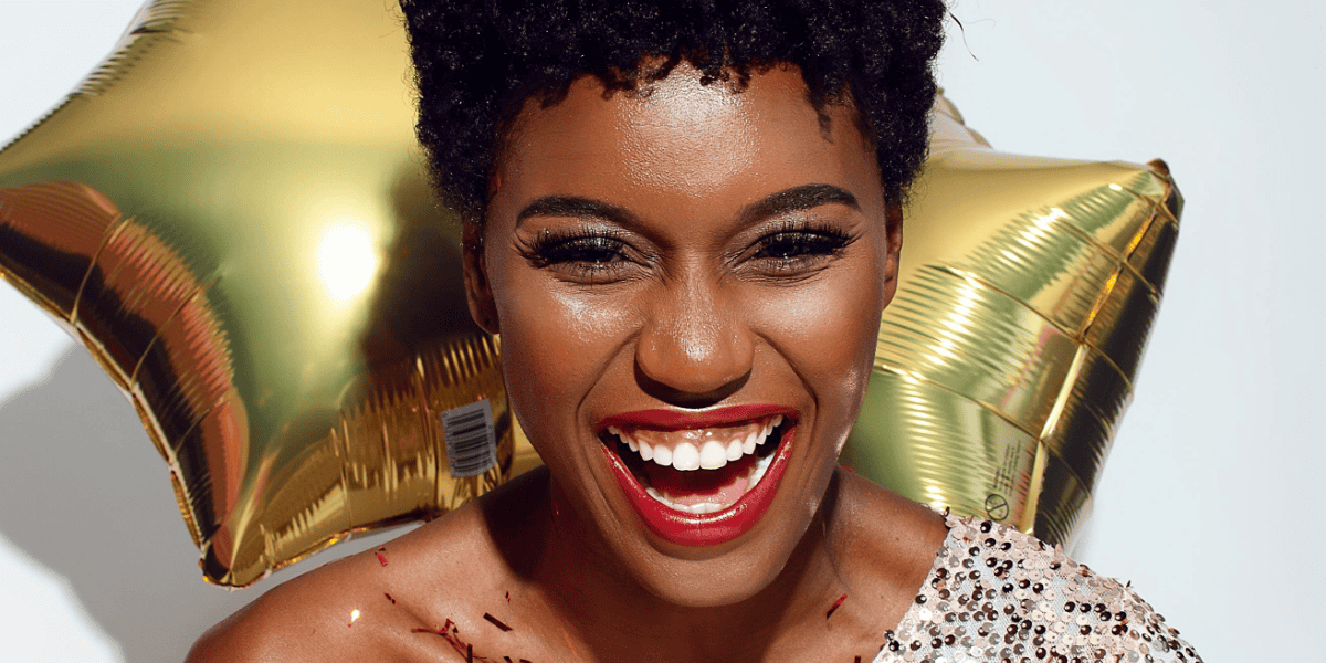A woman with vibrant makeup and glitter confetti on her shoulders, smiling brightly against a backdrop of golden balloons. This celebratory image embodies joy and positivity, aligning with the theme of "new year resolutions for perfectionists