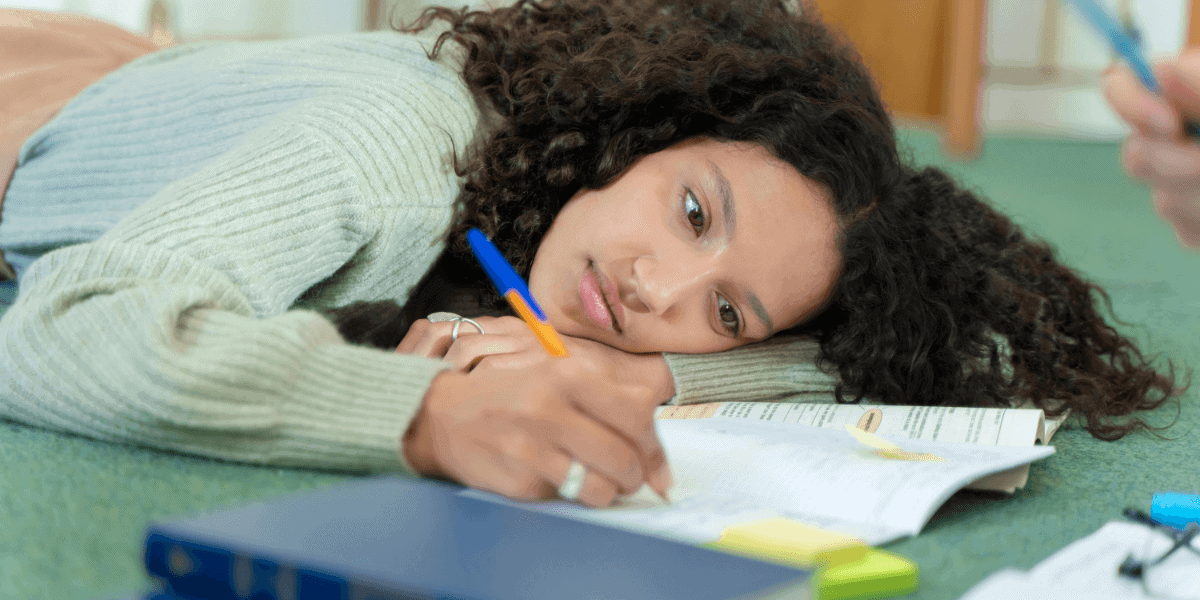 Young woman lying on the floor with her head resting on her arm, holding a pen and surrounded by open books and papers, reflecting a moment of distraction and lack of motivation, perfect for illustrating procrastination quotes