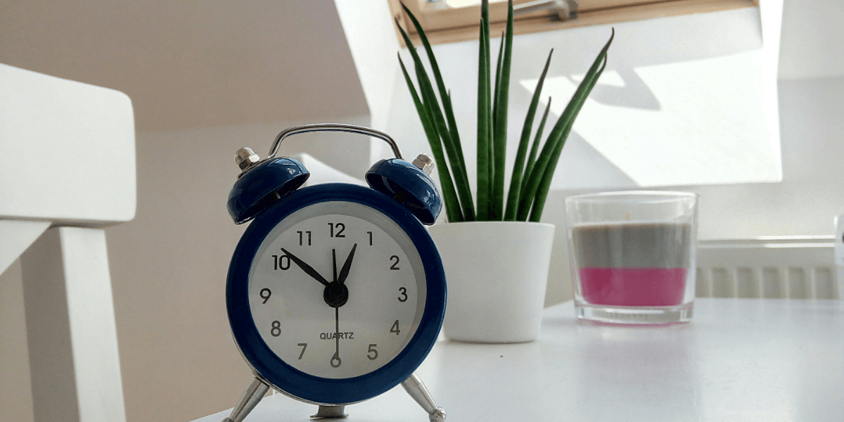 A blue alarm clock set at 10:10 on a white table, symbolizing productive things to do in a Pomodoro break, with a potted plant and a layered candle in the background