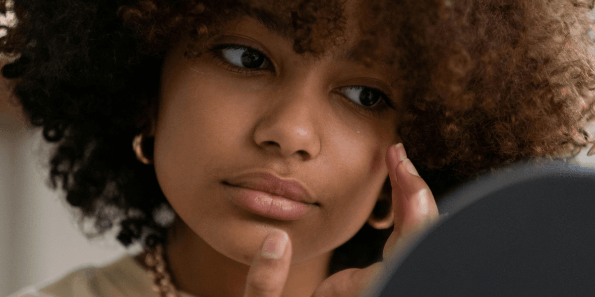 A young woman with curly hair thoughtfully looking in a mirror, accompanied by text reading '29 Healing February Journal Prompts for Self-Love'