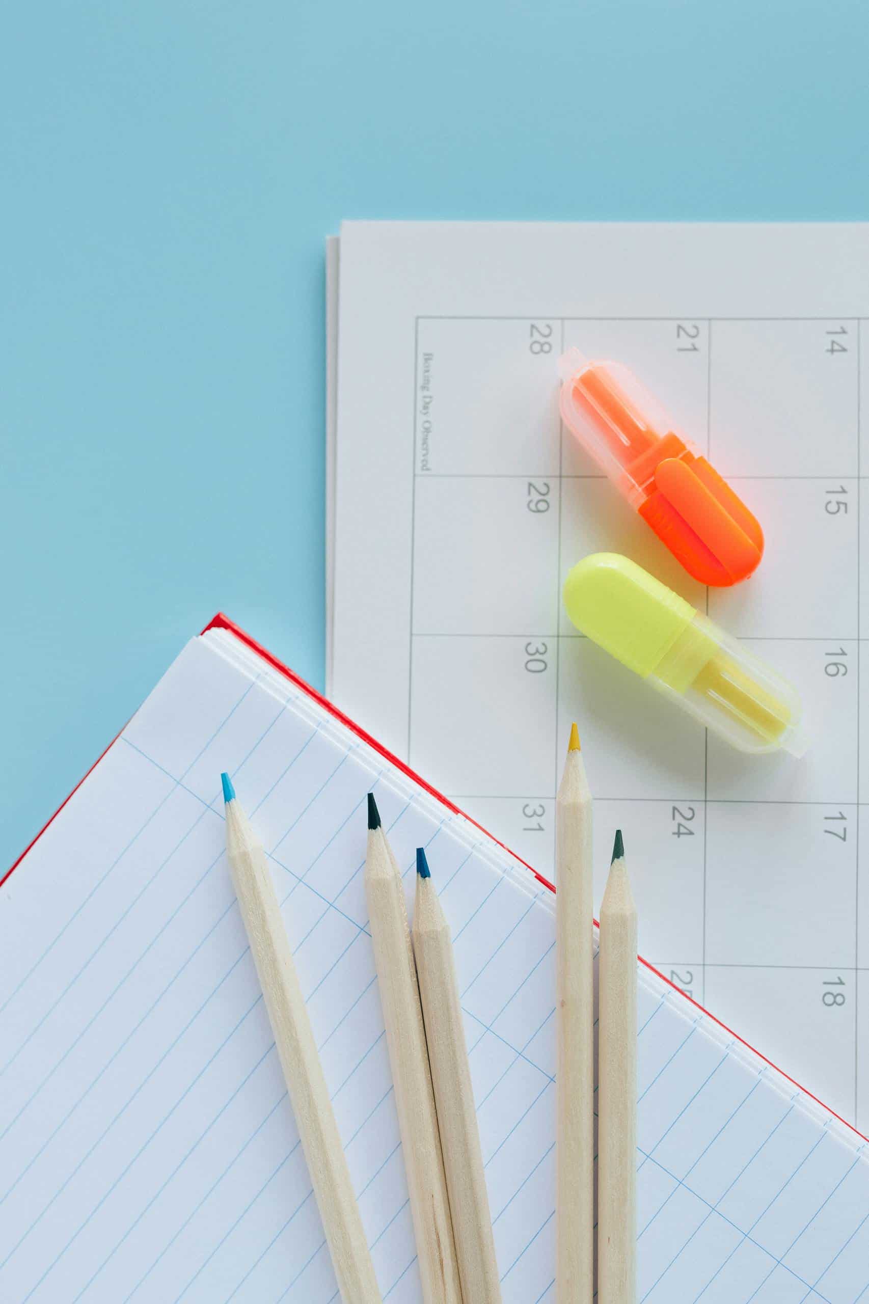 Overhead shot of calendar, notebook, wooden pencils, and highlighters on a blue background.